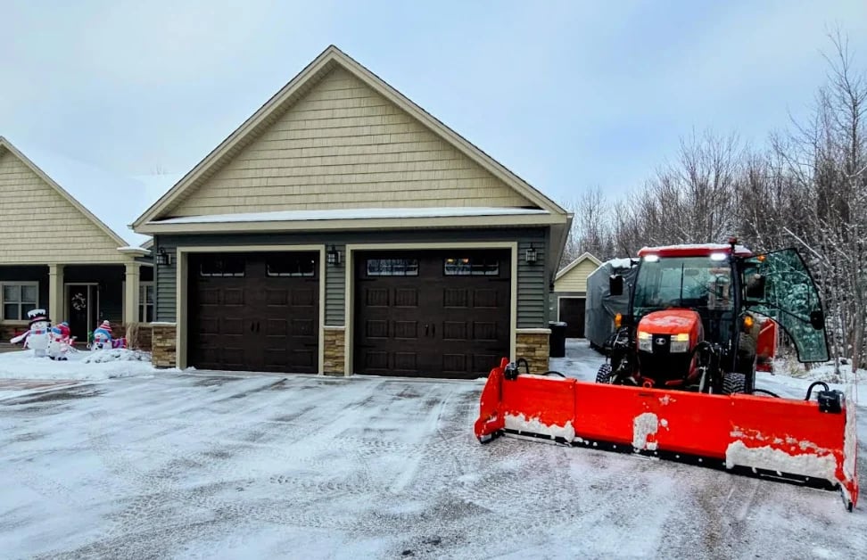 Red snow plow truck clearing driveway in front of two-car garage during winter
